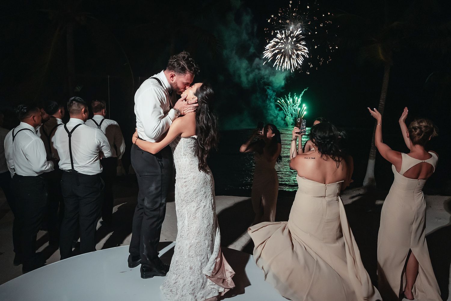 Bride and groom share first dance kiss with fireworks lighting the night sky at an outdoor wedding reception.