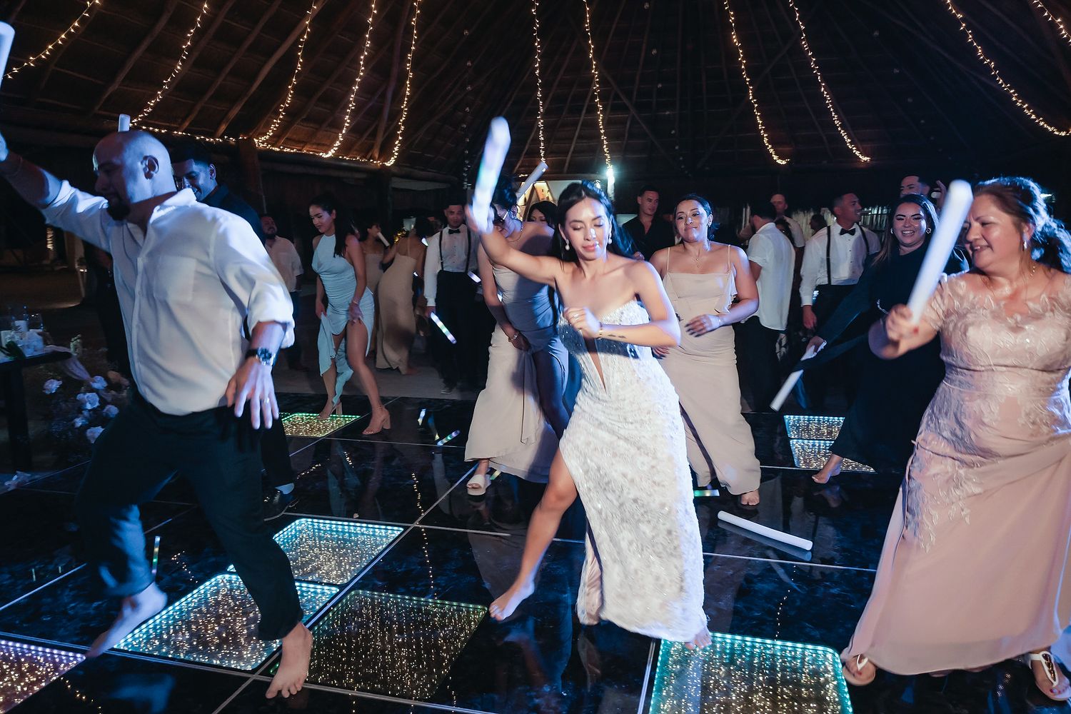 Bride dances energetically with guests holding glow sticks at a wedding reception under a tent with string lights.