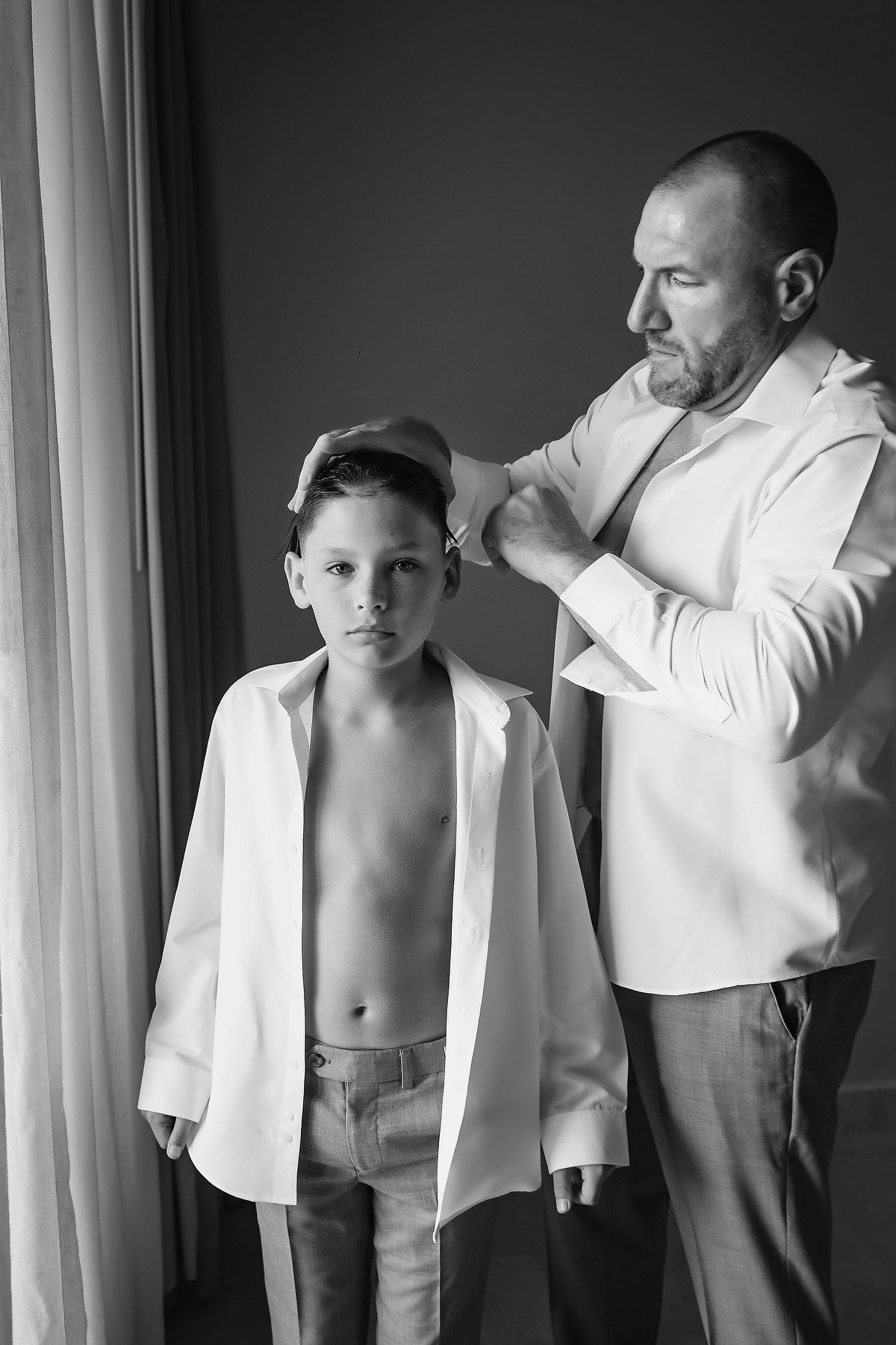 Black and white photo of a father helping his young son put on a white dress shirt, standing near a window.