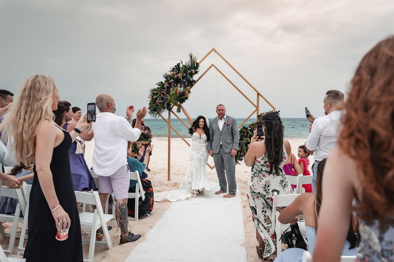 Couple walks down beach wedding aisle under geometric copper arch with floral decor, guests cheering on both sides.