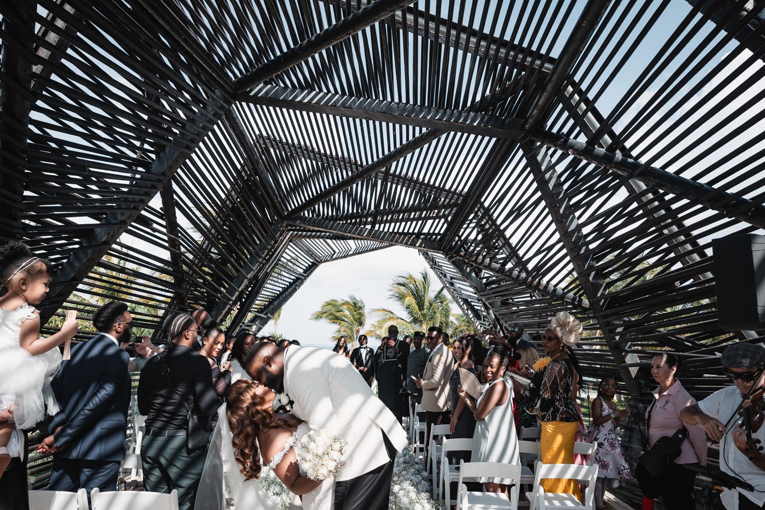 Guests applauding as bride and groom share first kiss under dramatic bamboo arch ceremony venue.