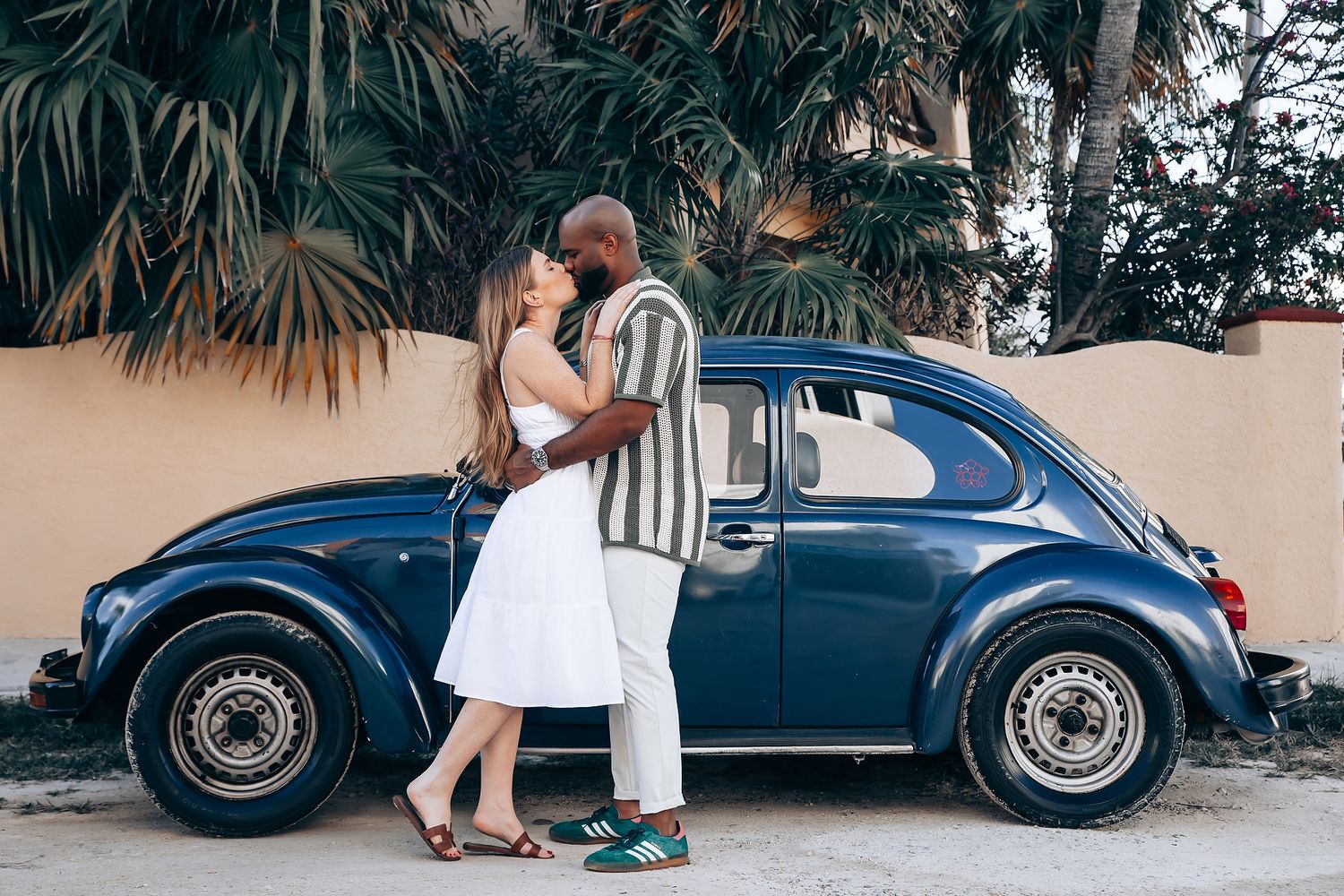 Interracial couple sharing a tender moment kissing beside a classic blue Volkswagen Beetle near palm trees.
