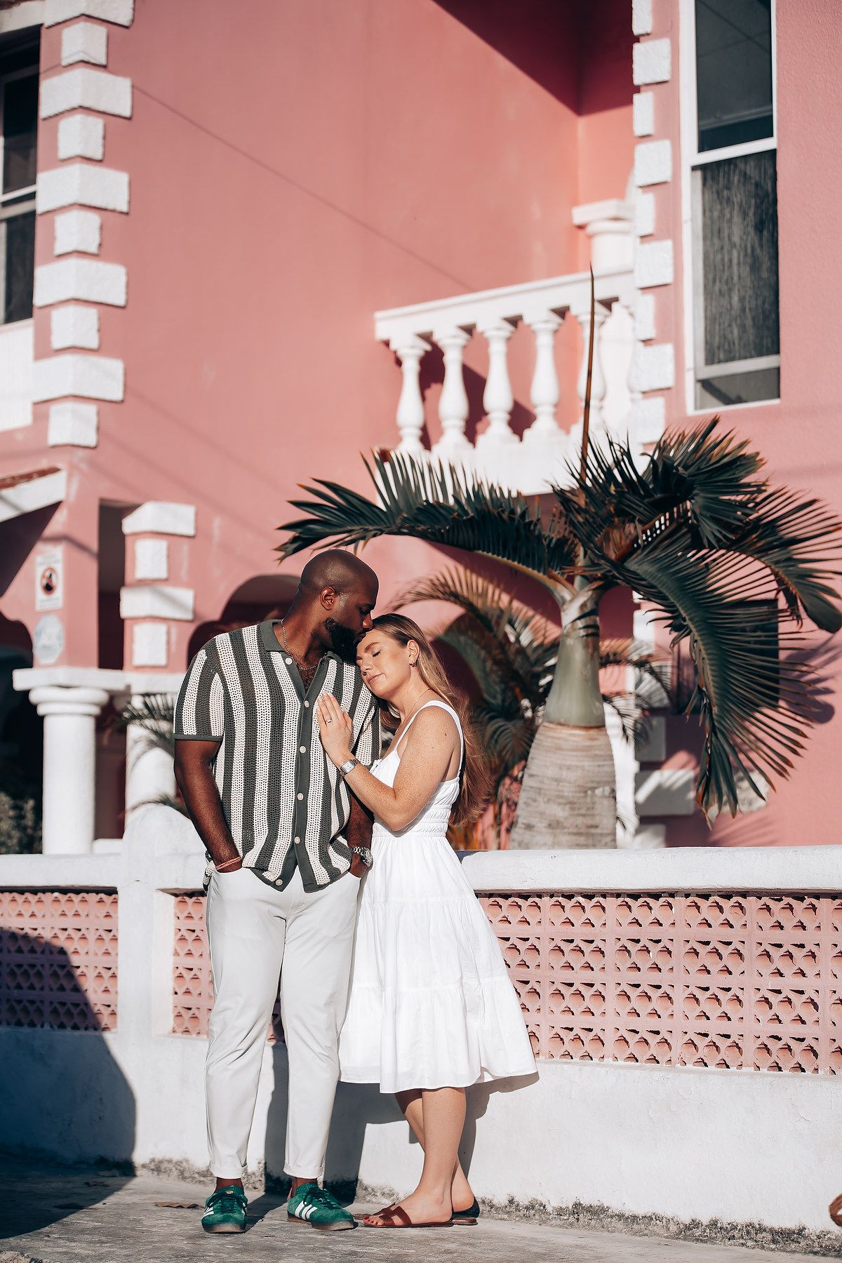 Couple sharing an intimate moment outside a vibrant pink colonial building with a tropical palm tree.