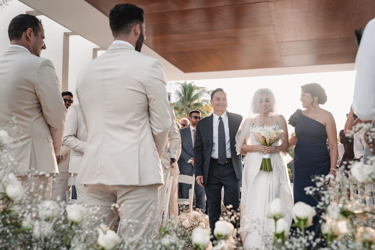 Bride walks down the aisle with escort at outdoor wedding ceremony surrounded by white floral arrangements.