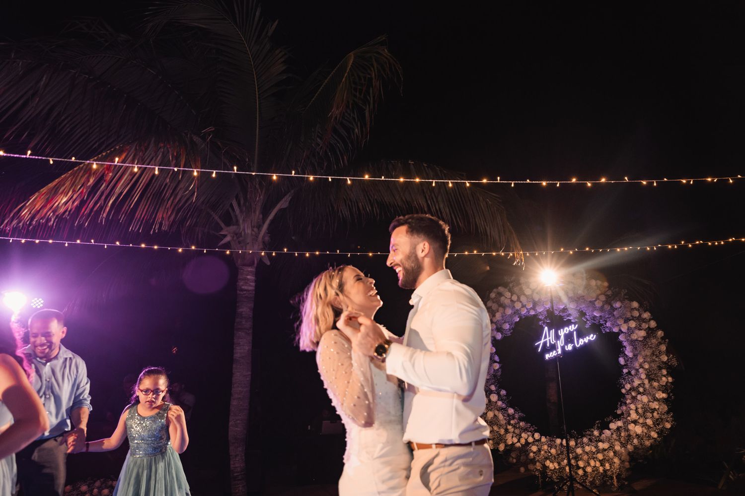 Bride and groom share romantic dance under string lights with neon sign backdrop at outdoor reception.