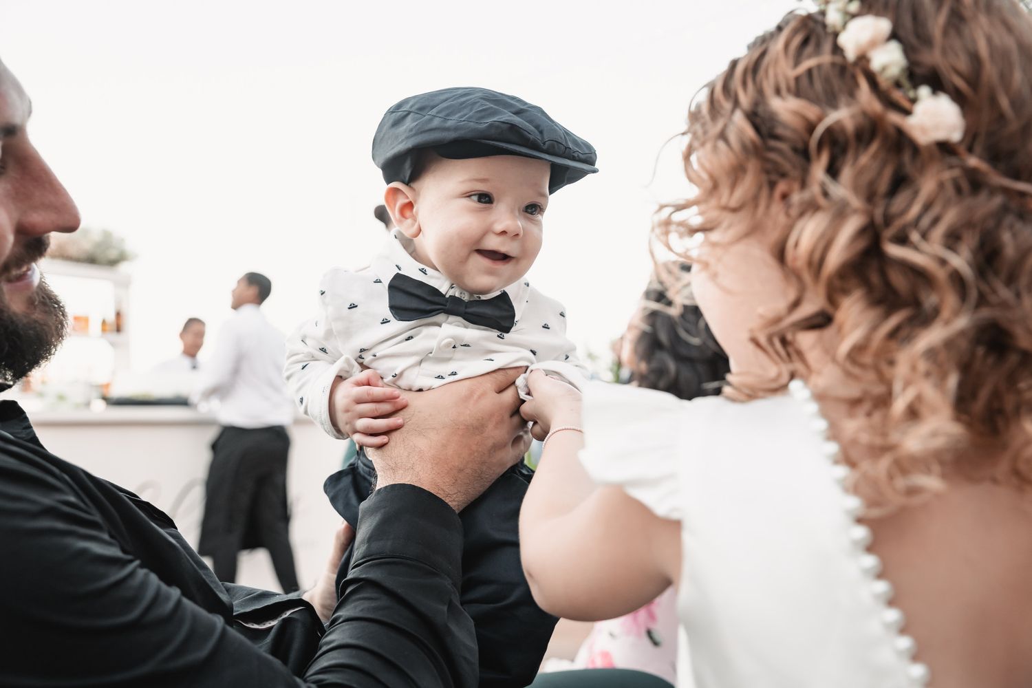 Smiling baby in a cap and bow tie held up by bride and groom at an outdoor wedding celebration.