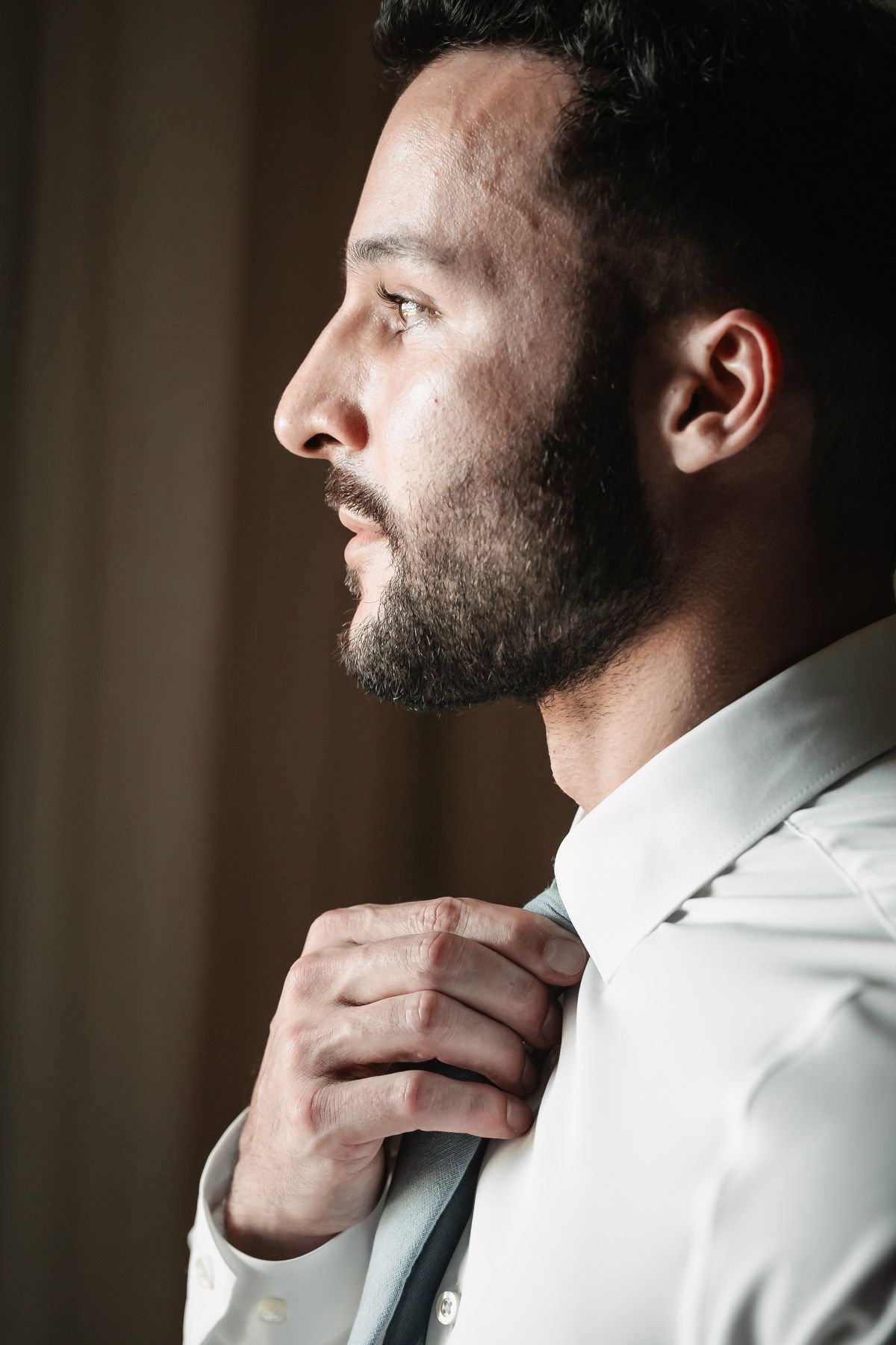 Groom in white shirt adjusting his tie in dramatic side profile lighting.