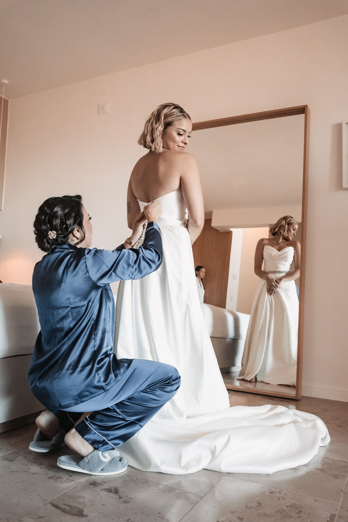 Bridesmaid kneeling to help bride into white wedding gown reflected in a large mirror.