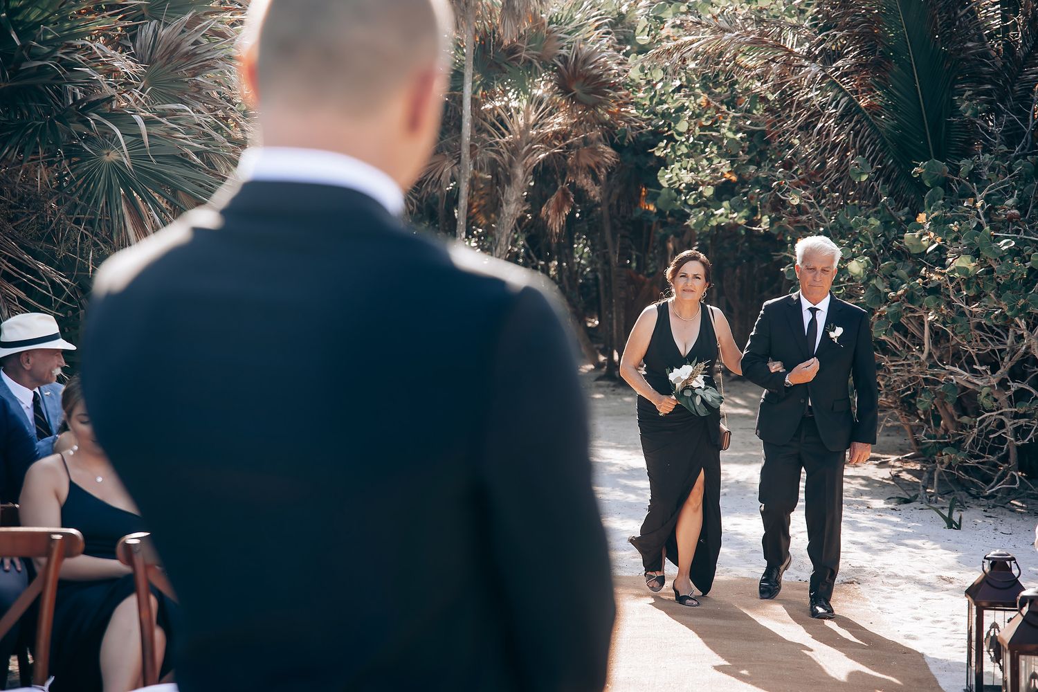 Wedding ceremony guests gather on a garden path lined with wooden posts and tropical foliage.