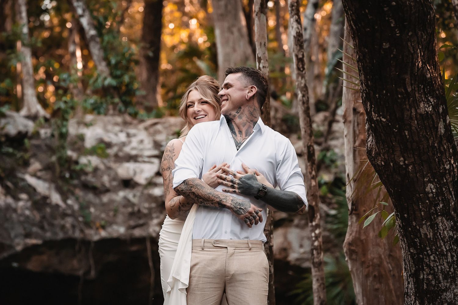 Couple with tattoos embracing in a lush forest setting, smiling warmly during an outdoor engagement photo session.