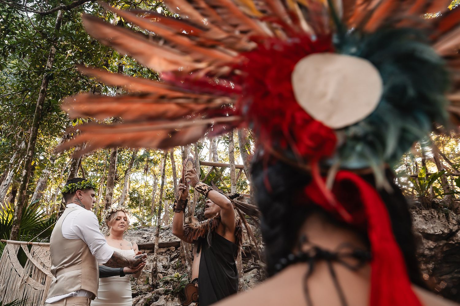 Mayan ceremony officiant in colorful feathered headdress performs traditional wedding ritual outdoors in lush jungle setting.