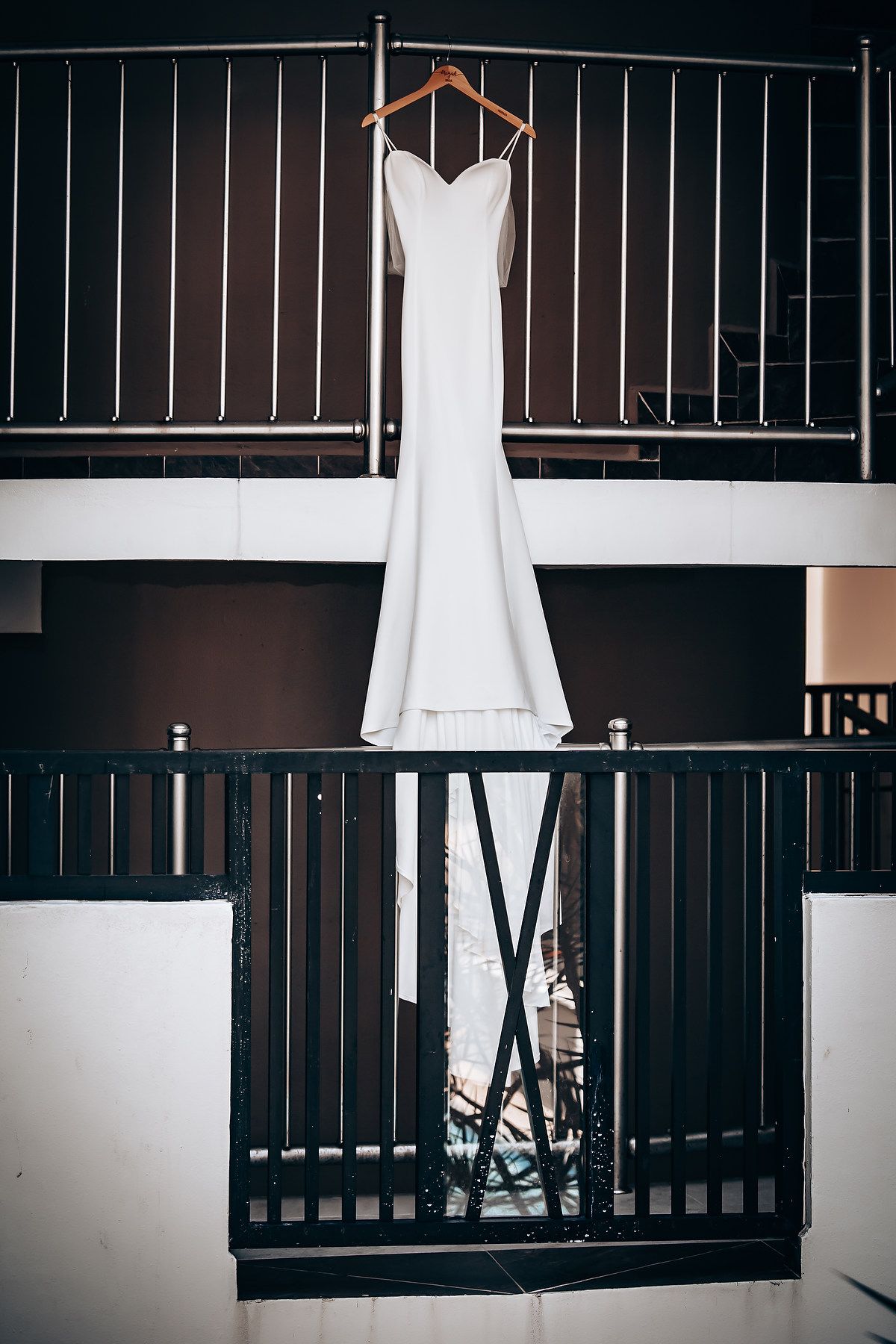 An elegant white wedding dress hangs against a dark wooden railing in natural light.