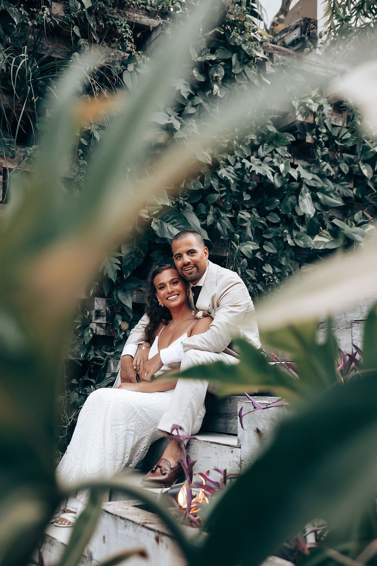 Couple in white attire pose together among lush green foliage in a romantic garden setting.