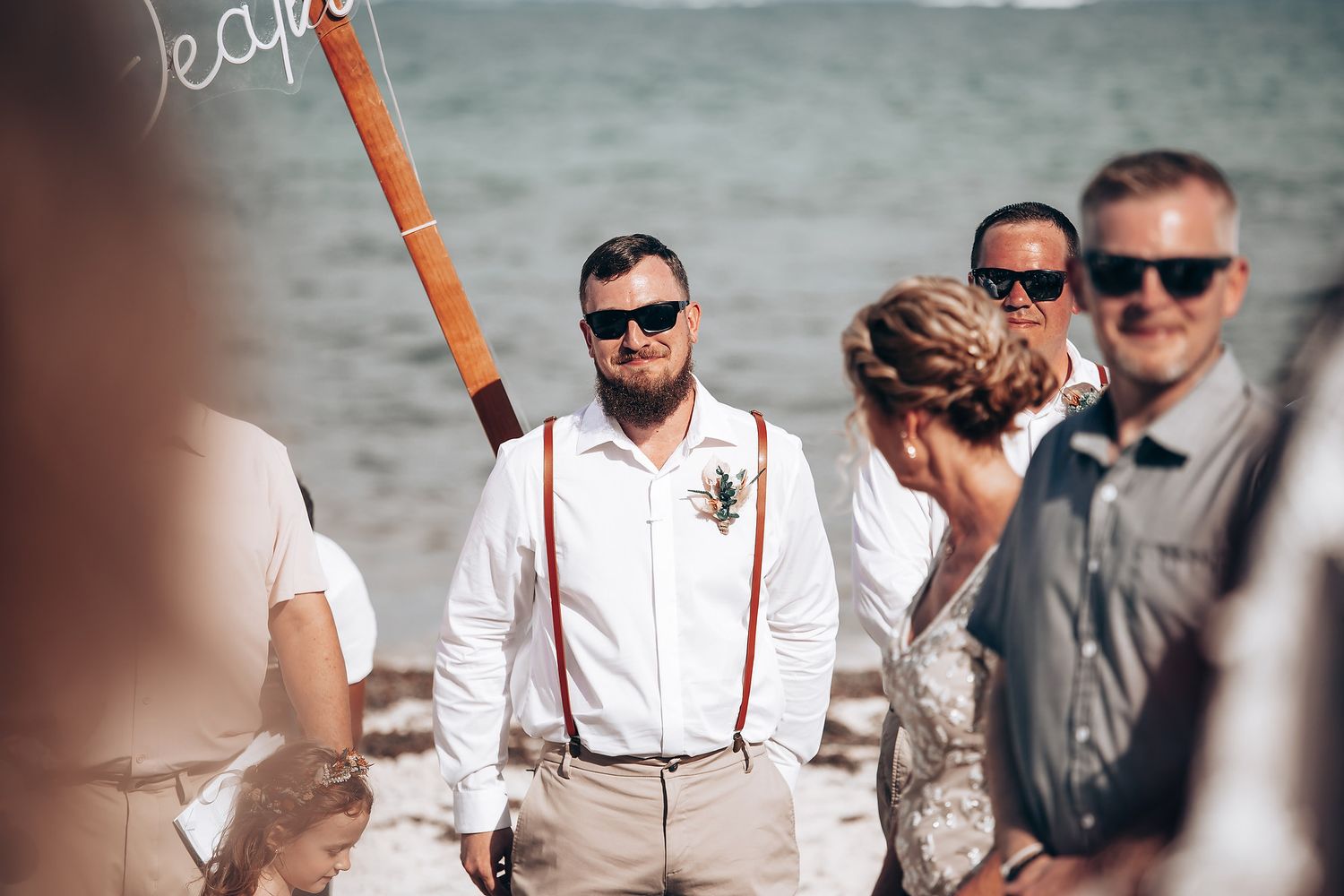Wedding party in white and khaki attire walking along beach with ocean backdrop.