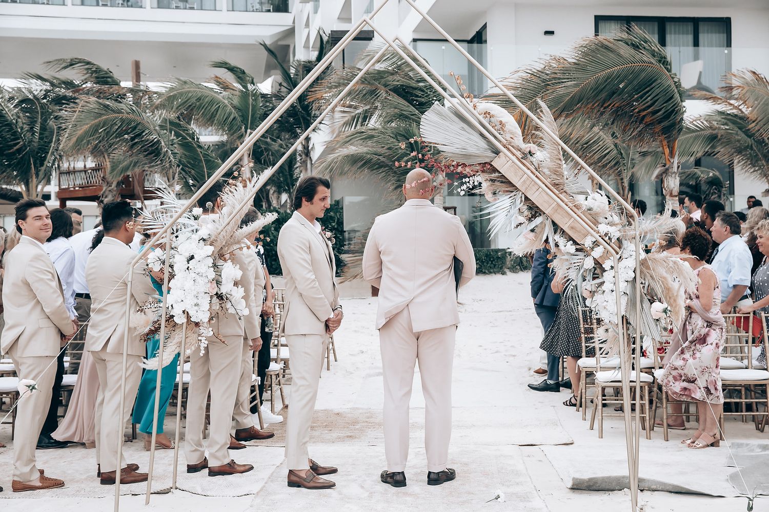 Guests seated at a beachside wedding ceremony under white decorative arches and palm trees.