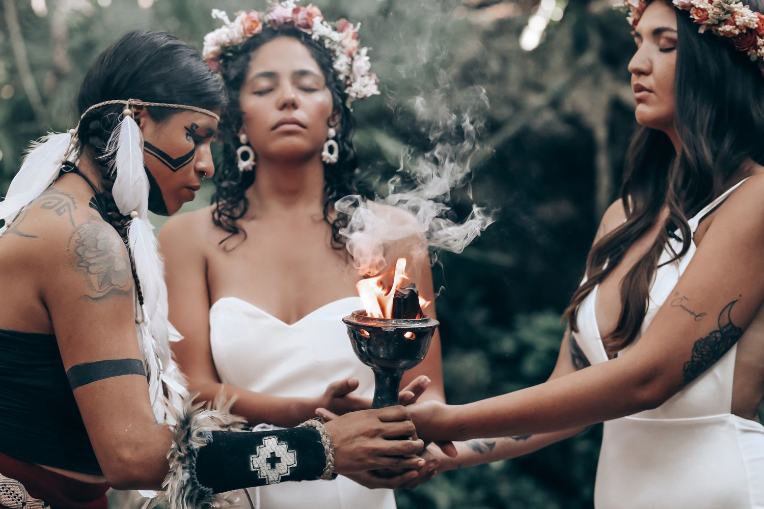 Couple wearing white dresses and floral crowns share a tender moment in a forest setting.