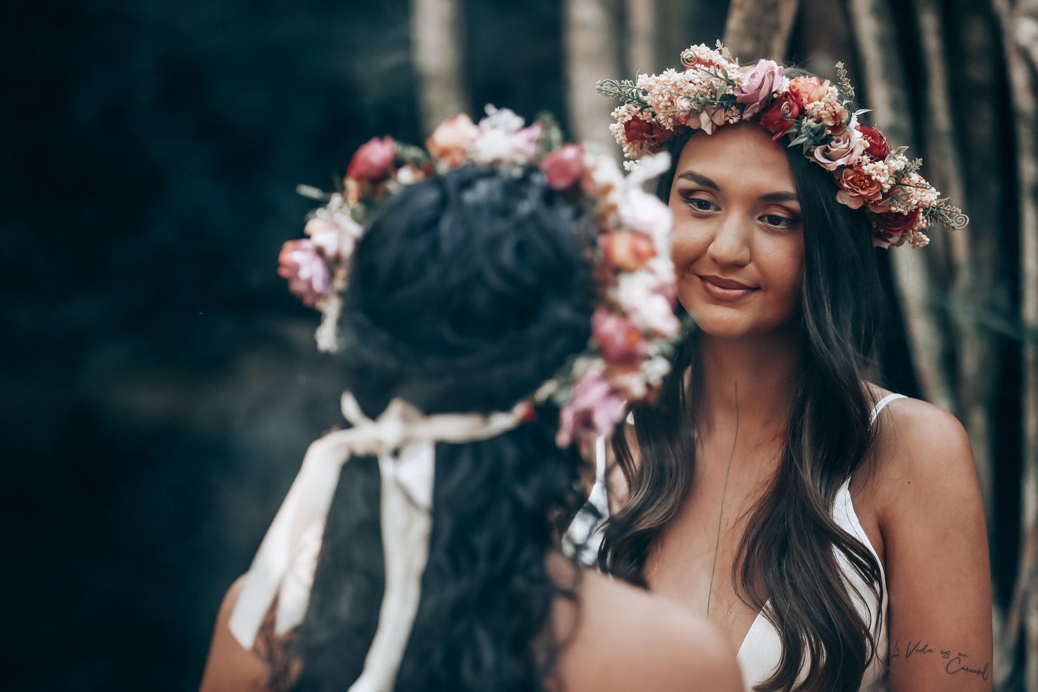 Two people in white dresses wearing floral crowns embrace in a tropical forest setting.