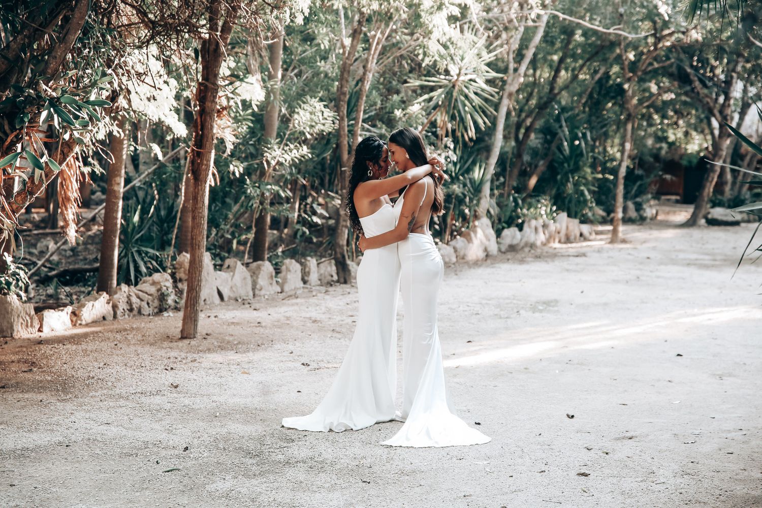 Two figures in white attire embrace in a romantic outdoor setting with lush greenery and trees in the background.