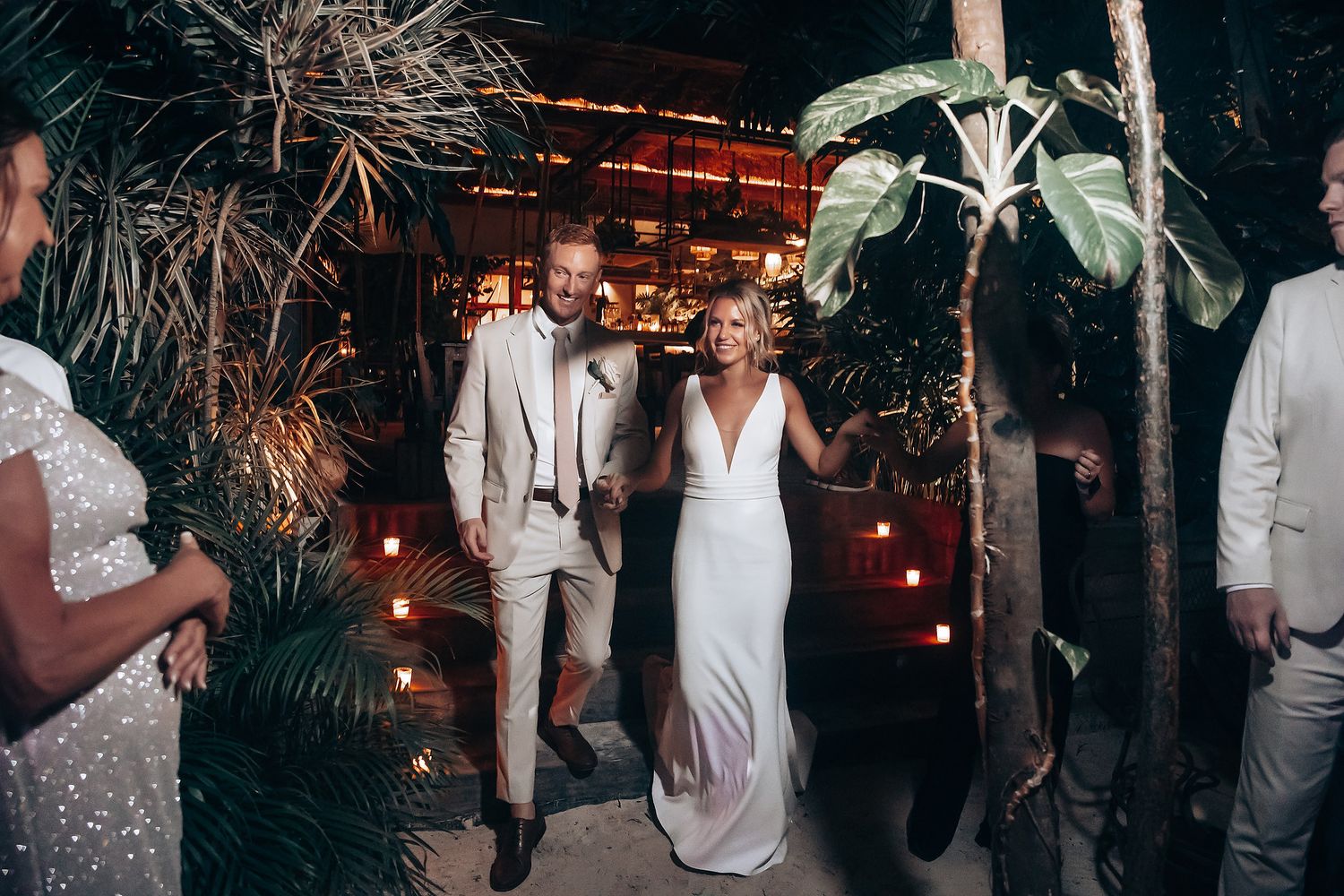 A romantic wedding couple dances together under palm trees and string lights at an evening reception.