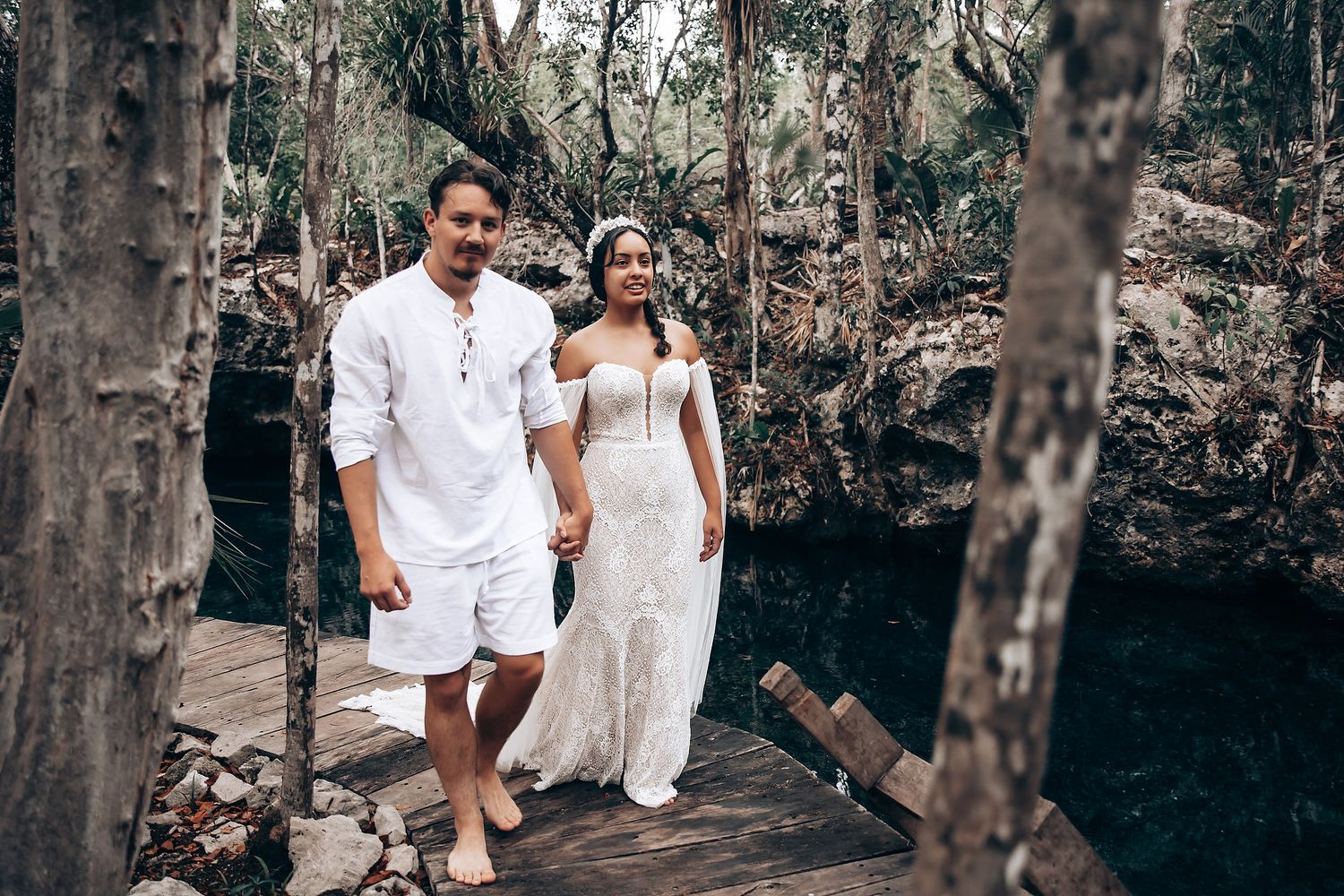 A couple in white outfits walks through a wooded area during a romantic photoshoot.