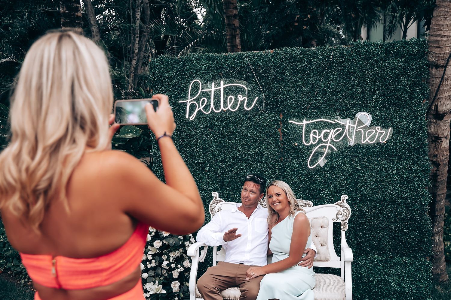 Outdoor wedding reception setup with greenery wall and white furniture.