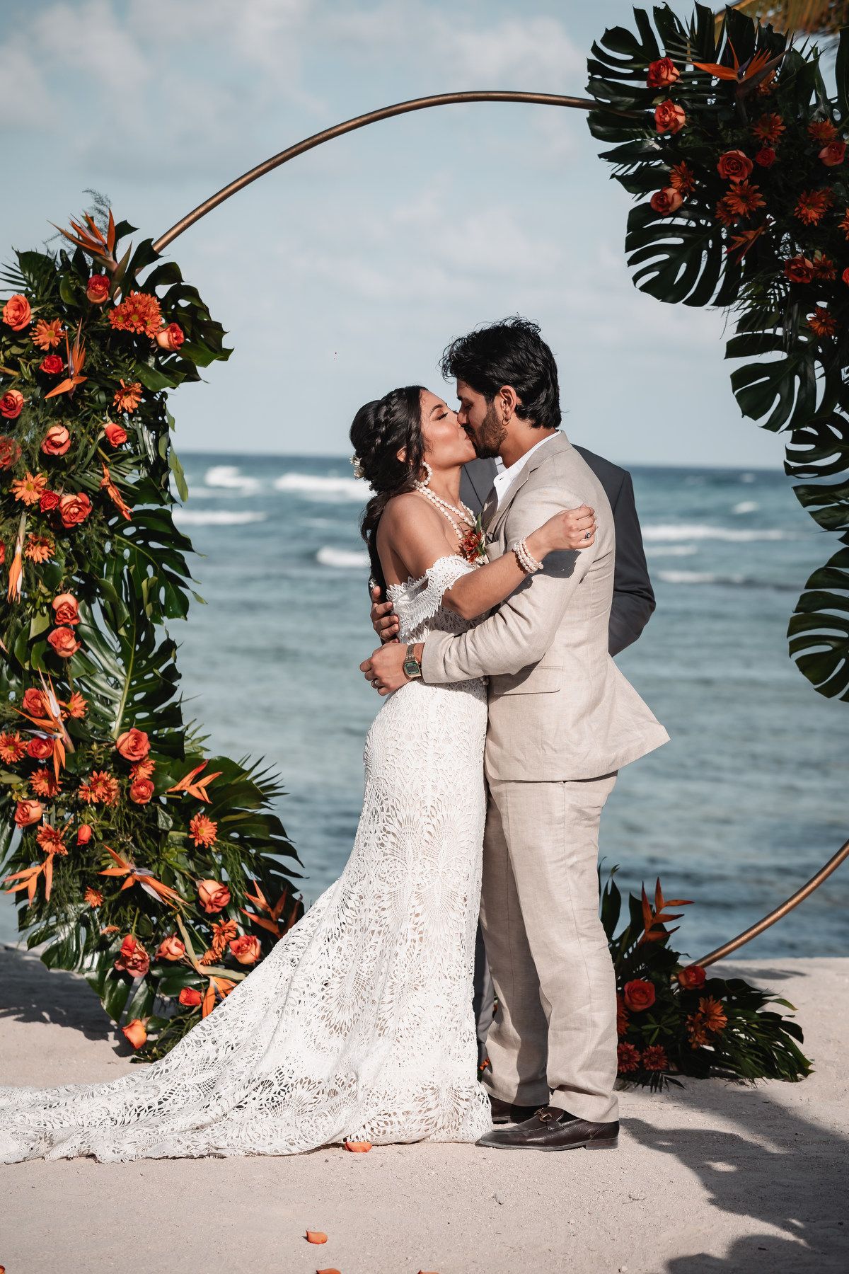 Bride and groom share first kiss under circular floral arch with ocean backdrop on sandy beach.