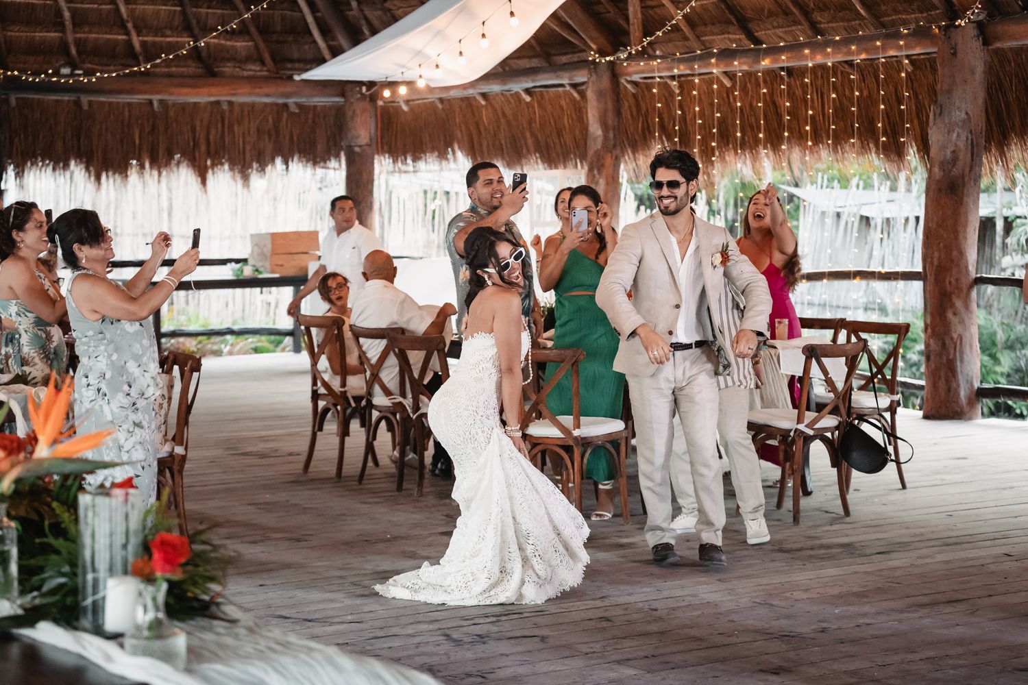 Bride and groom making grand entrance at tropical open-air reception venue with string lights and guests applauding.