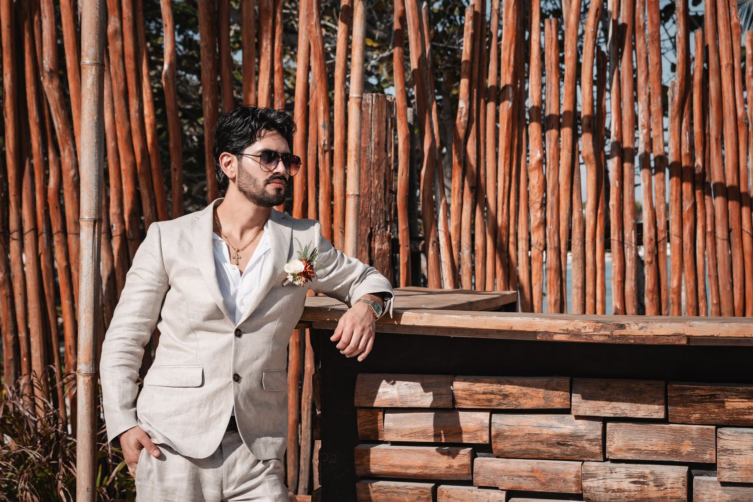 Elegant groom in tan linen suit leaning against rustic bamboo fence wall outdoors.