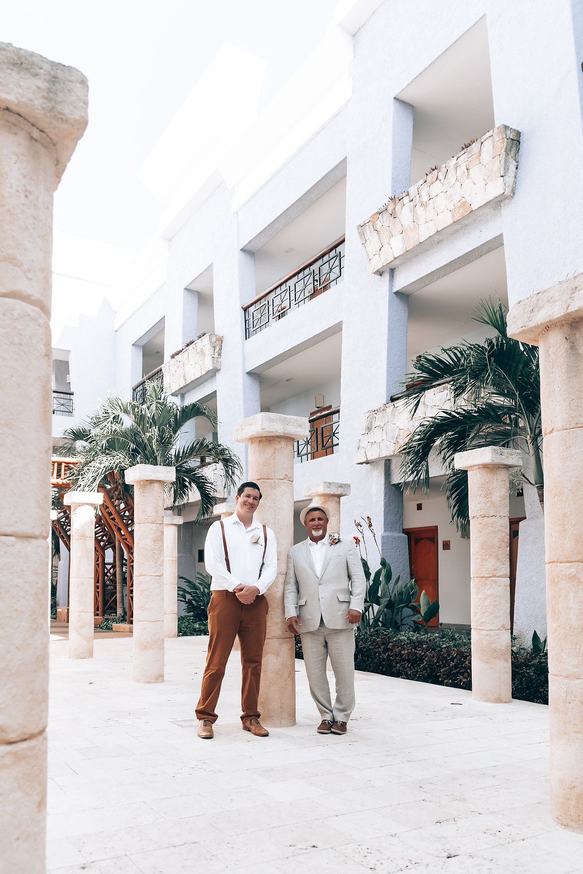 A bright white colonnade walkway with palm trees features two people walking in formal attire.