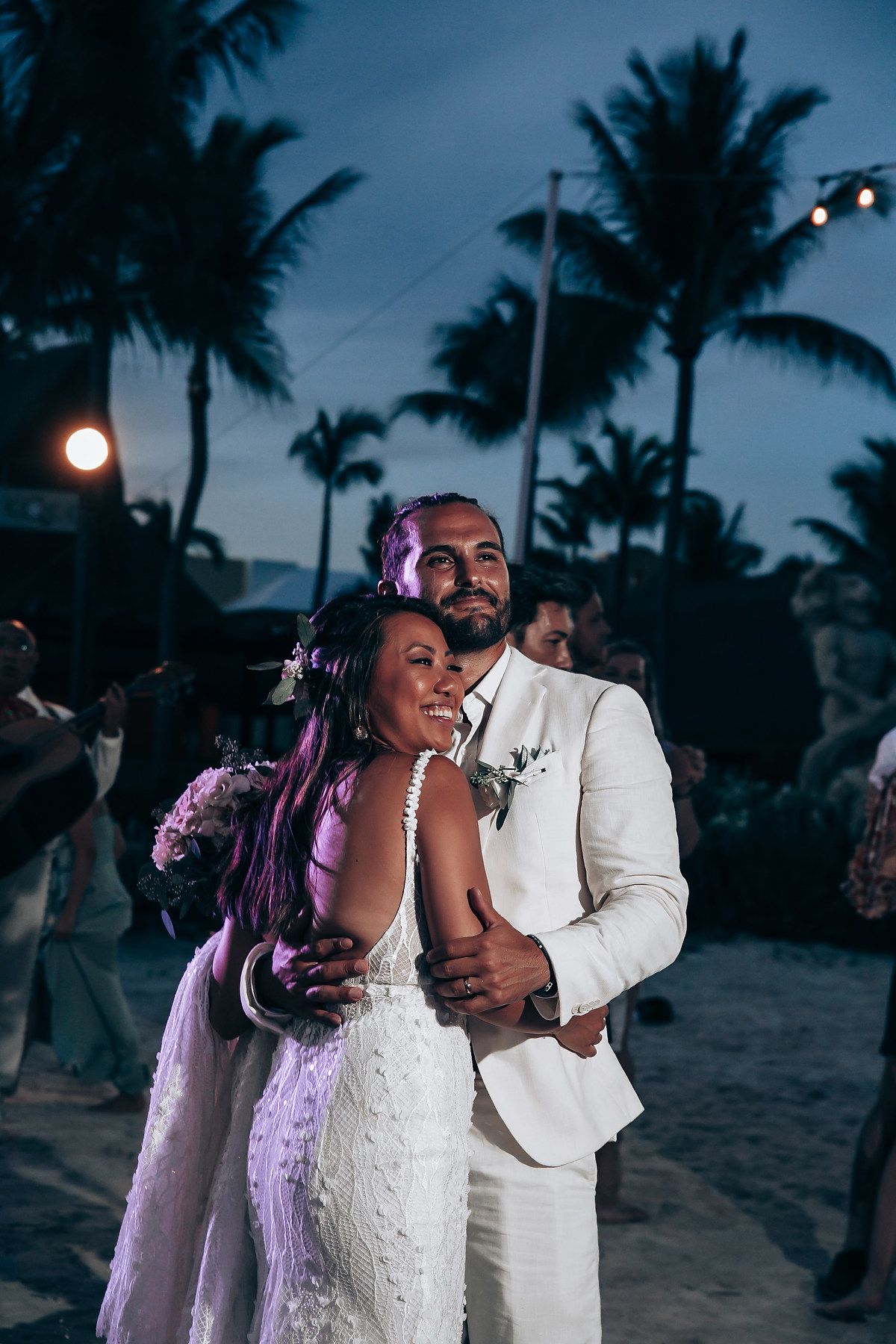 Couple dancing at twilight beach wedding reception under palm trees and purple lighting.
