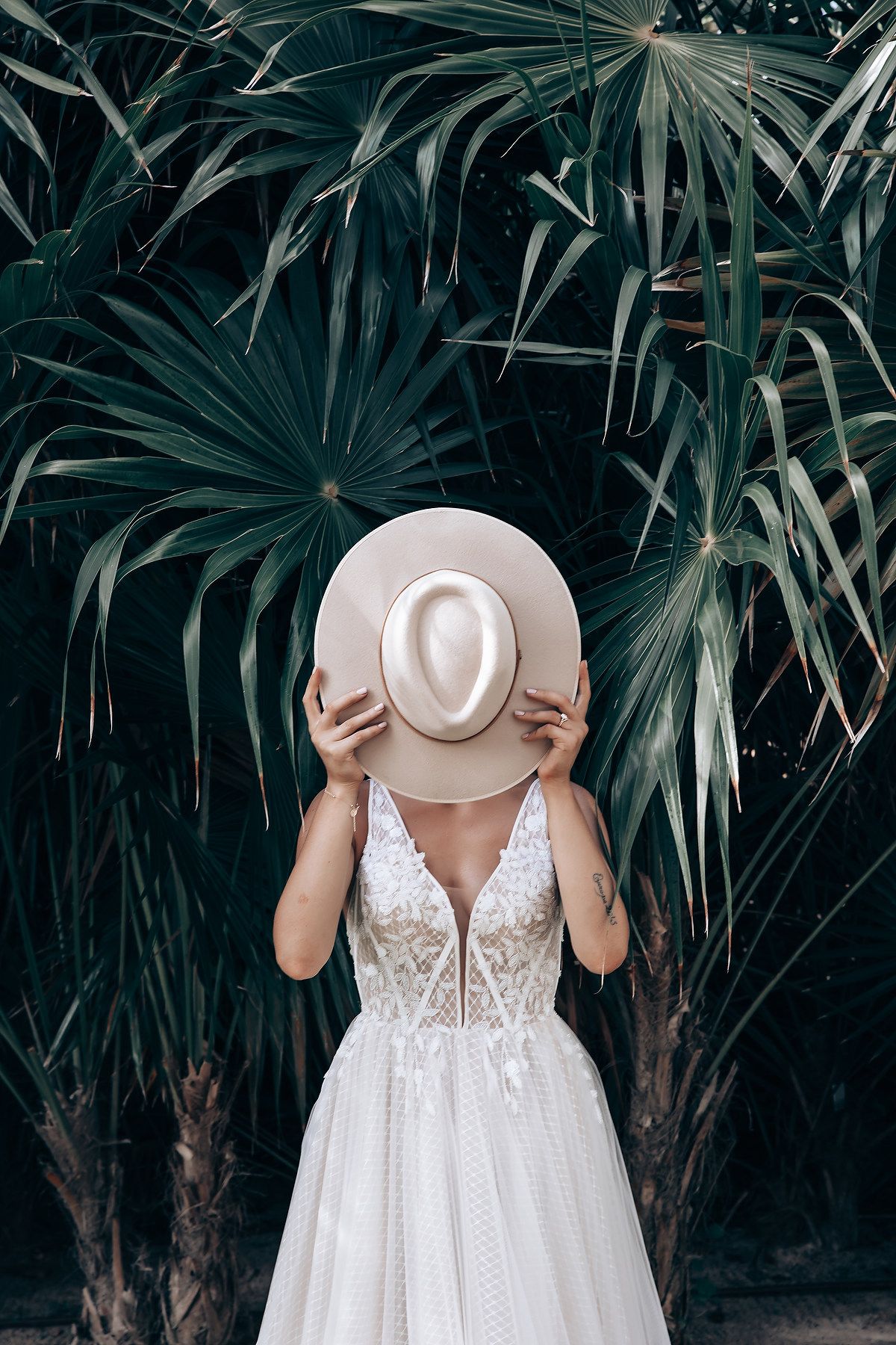 A person in a white lace wedding dress holds a cream-colored hat in front of tropical palm leaves.