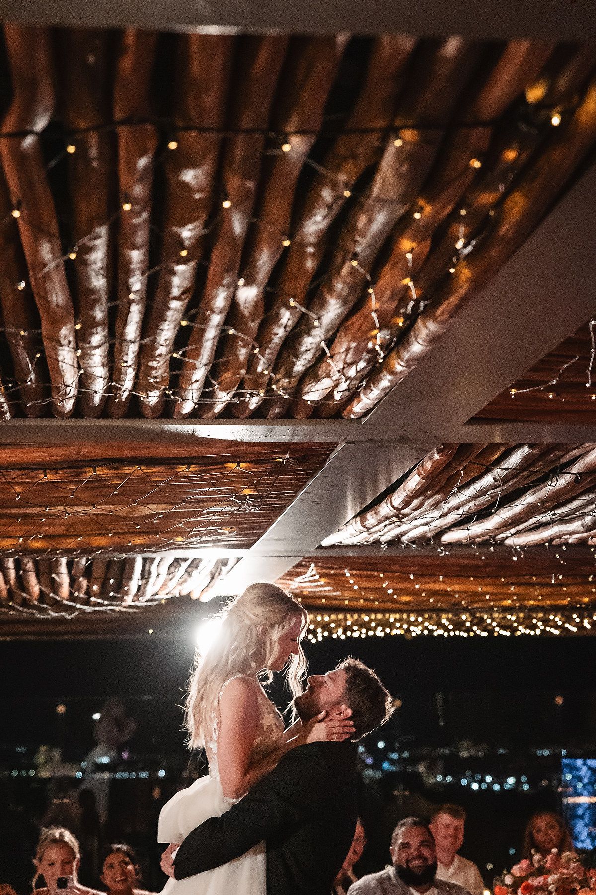 Wedding reception venue features exposed wooden beams with string lights creating a warm ambiance.