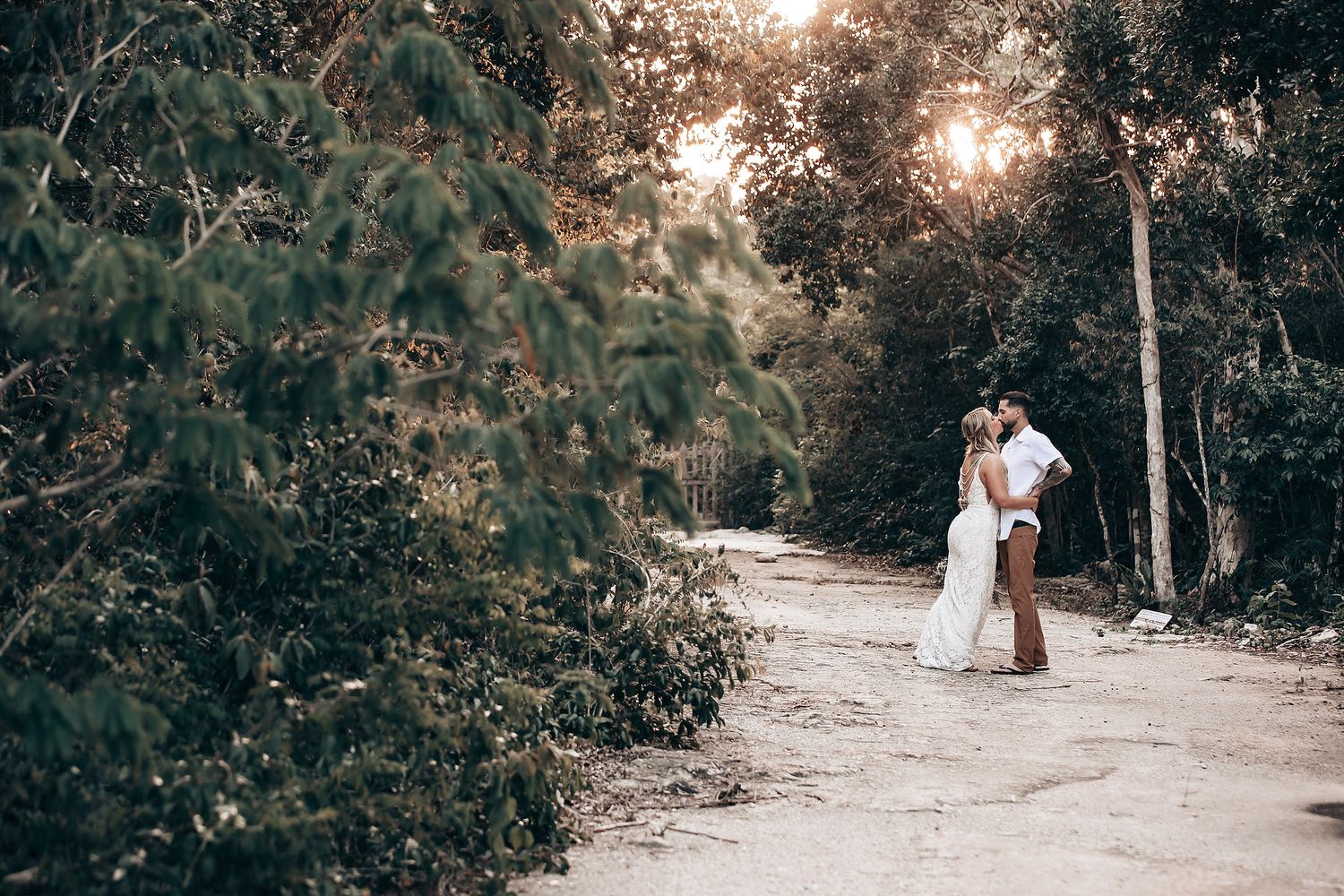 A romantic outdoor wedding photoshoot in a sunlit wooded path with natural greenery and trees.