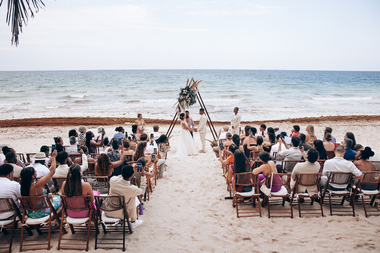 Wedding guests seated in white chairs on a tropical beach during an outdoor ceremony.