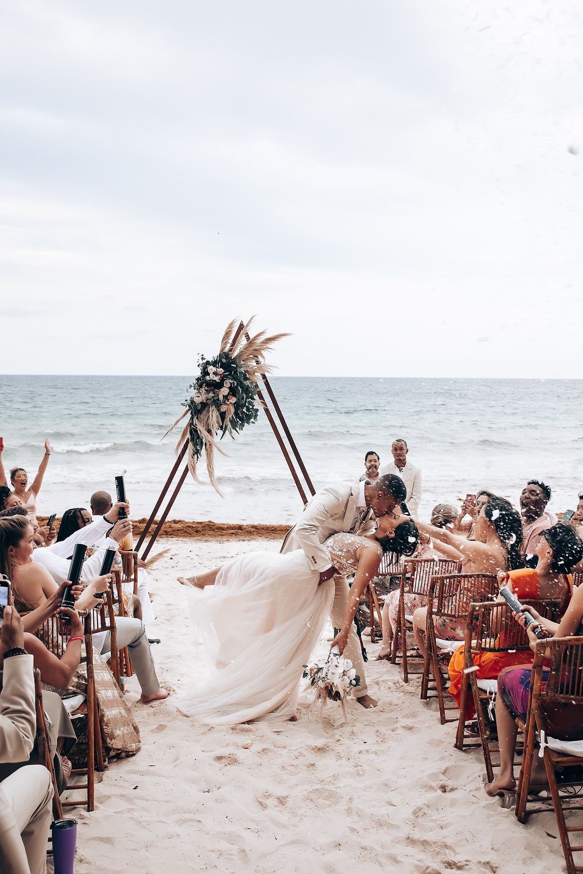 A joyous beach wedding celebration under a wooden arch decorated with pampas grass and flowers.