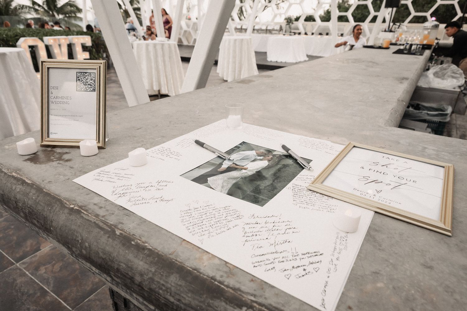 Wedding guest book display with signatures, framed photo, and elegant white decor at outdoor reception venue.