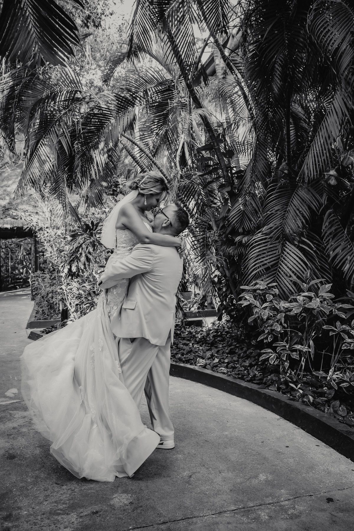 Groom lifts bride in joyful embrace surrounded by tropical palm trees in black and white photo.