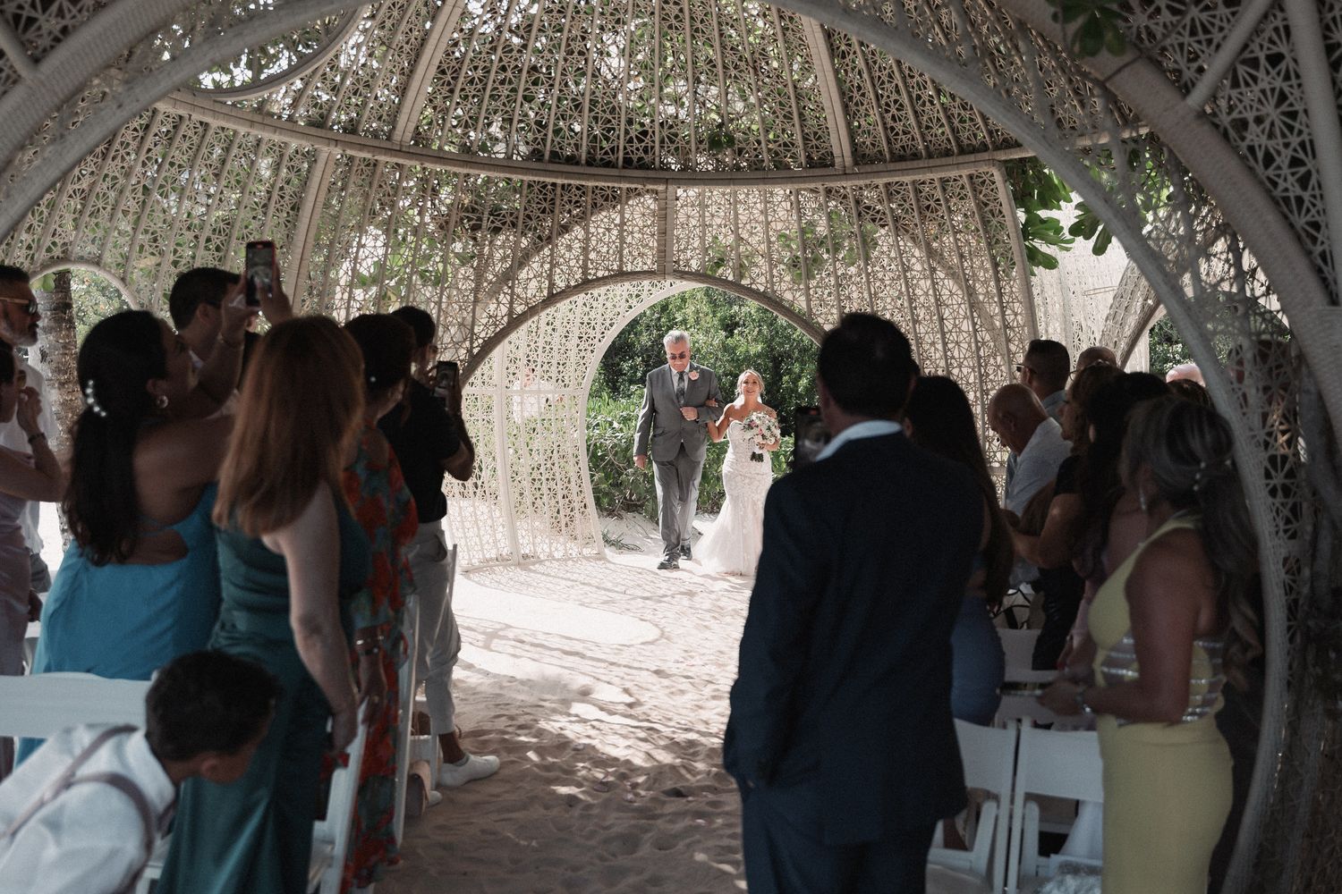 Wedding processional with bride and escort walking down aisle under ornate arch with seated guests.