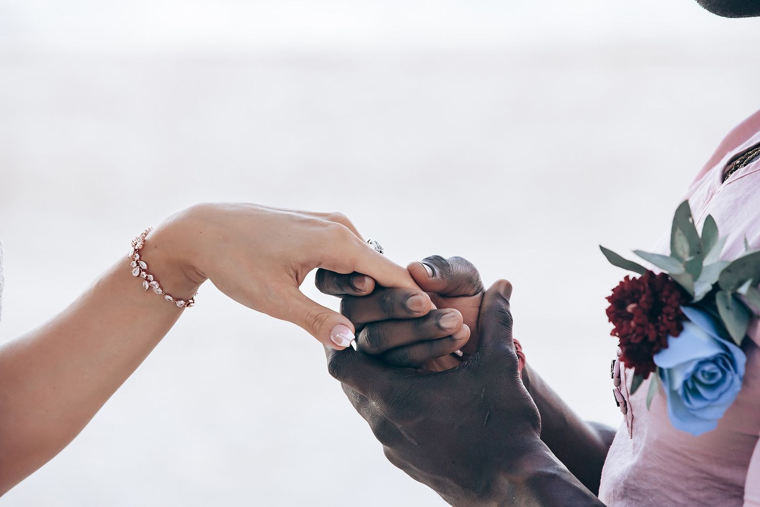 A couple holds hands as they walk on a beach, surrounded by guests in formal attire.