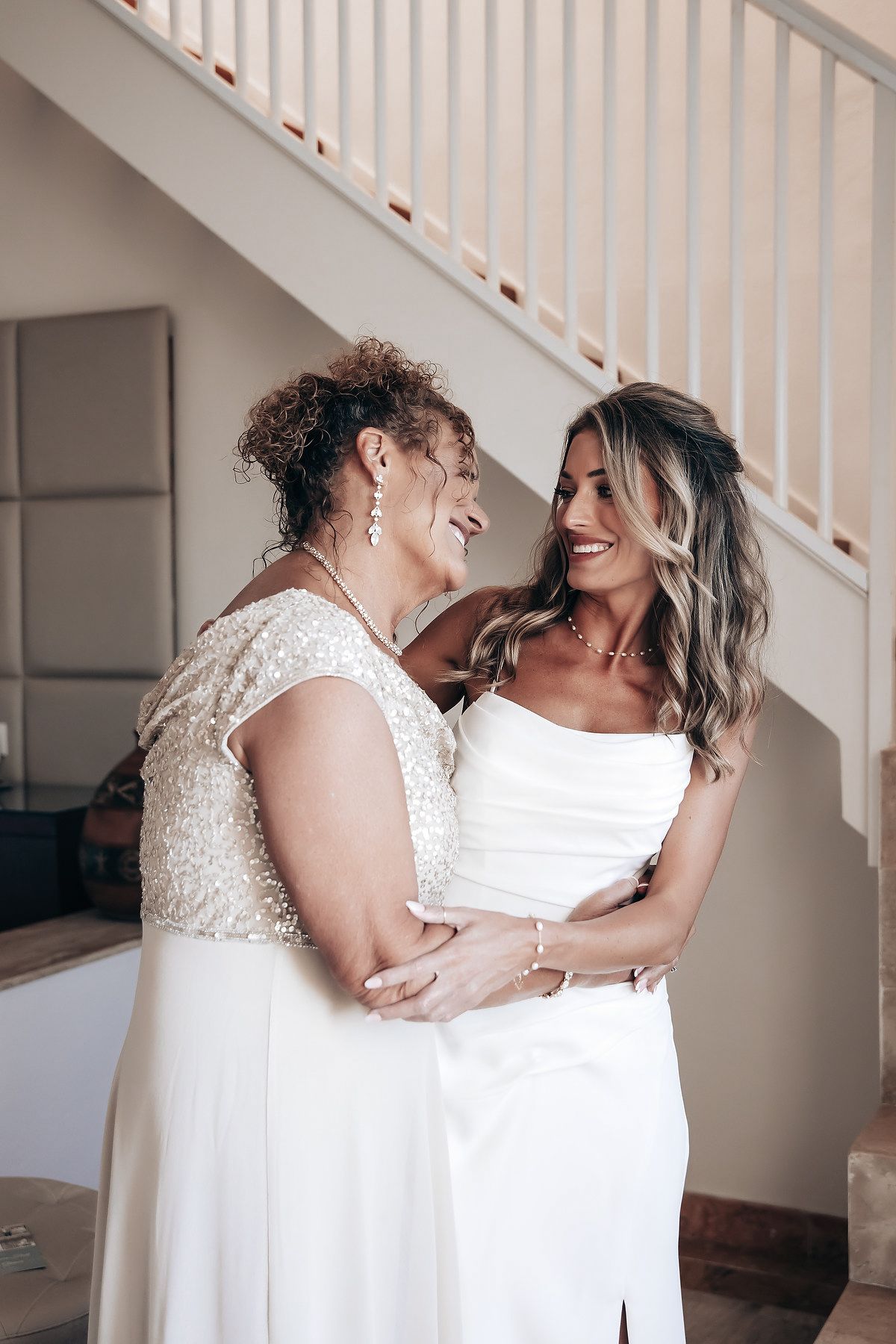 Two women in white wedding dresses embrace and smile at each other.