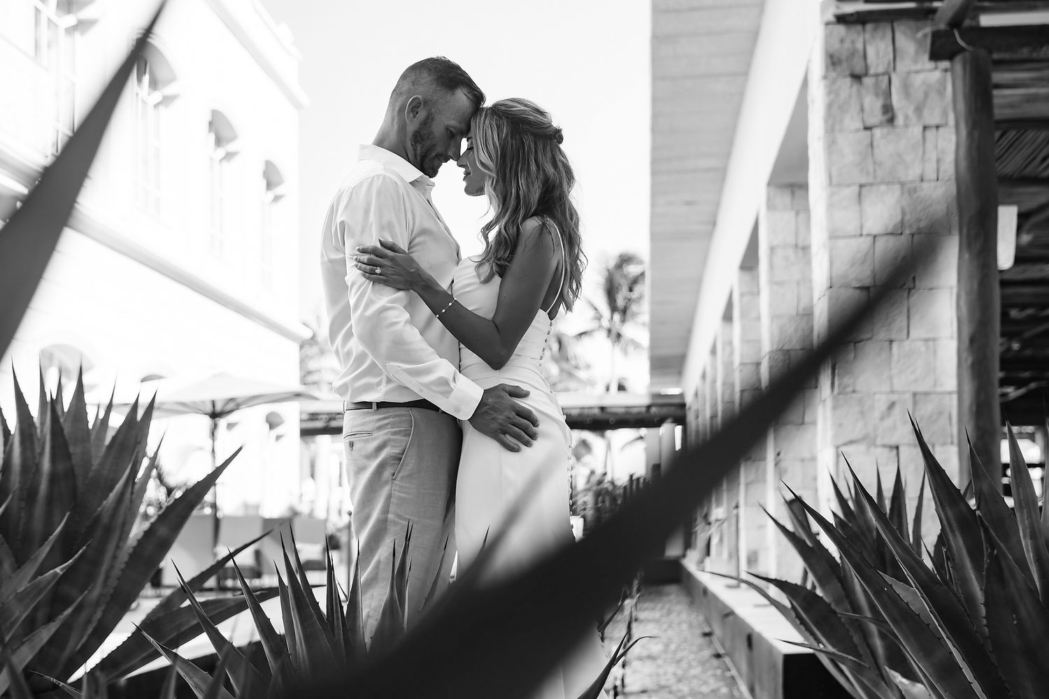 A black and white image of a couple embracing and gazing at each other in an outdoor setting with plants and foliage in the background.