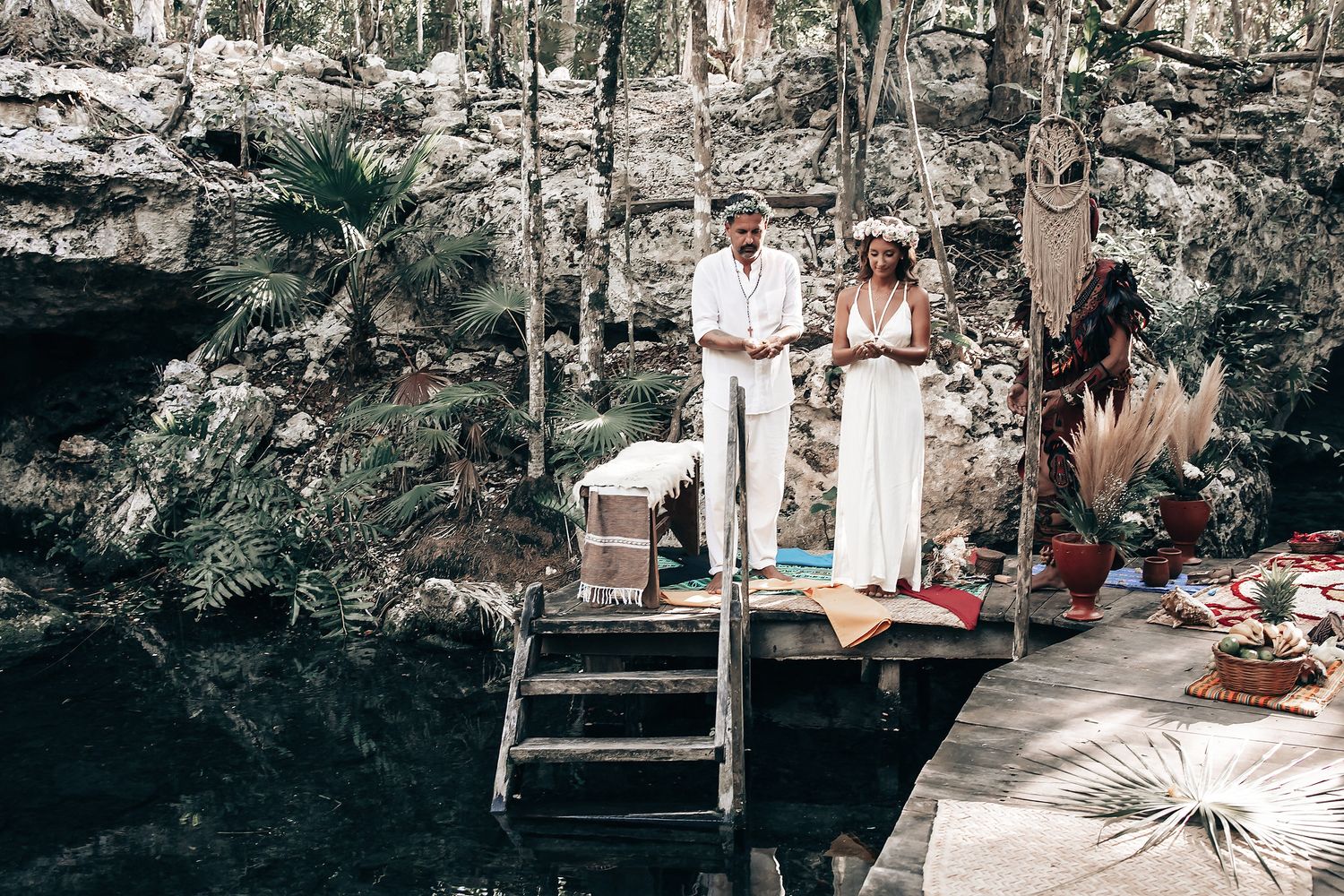A couple standing on a wooden platform in a lush forest with a body of water in the background.