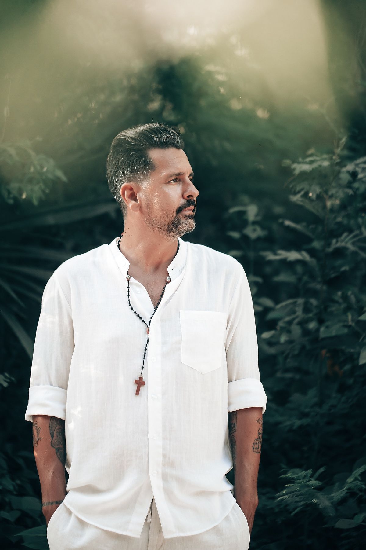 A man in white clothing stands in a lush, forested setting.