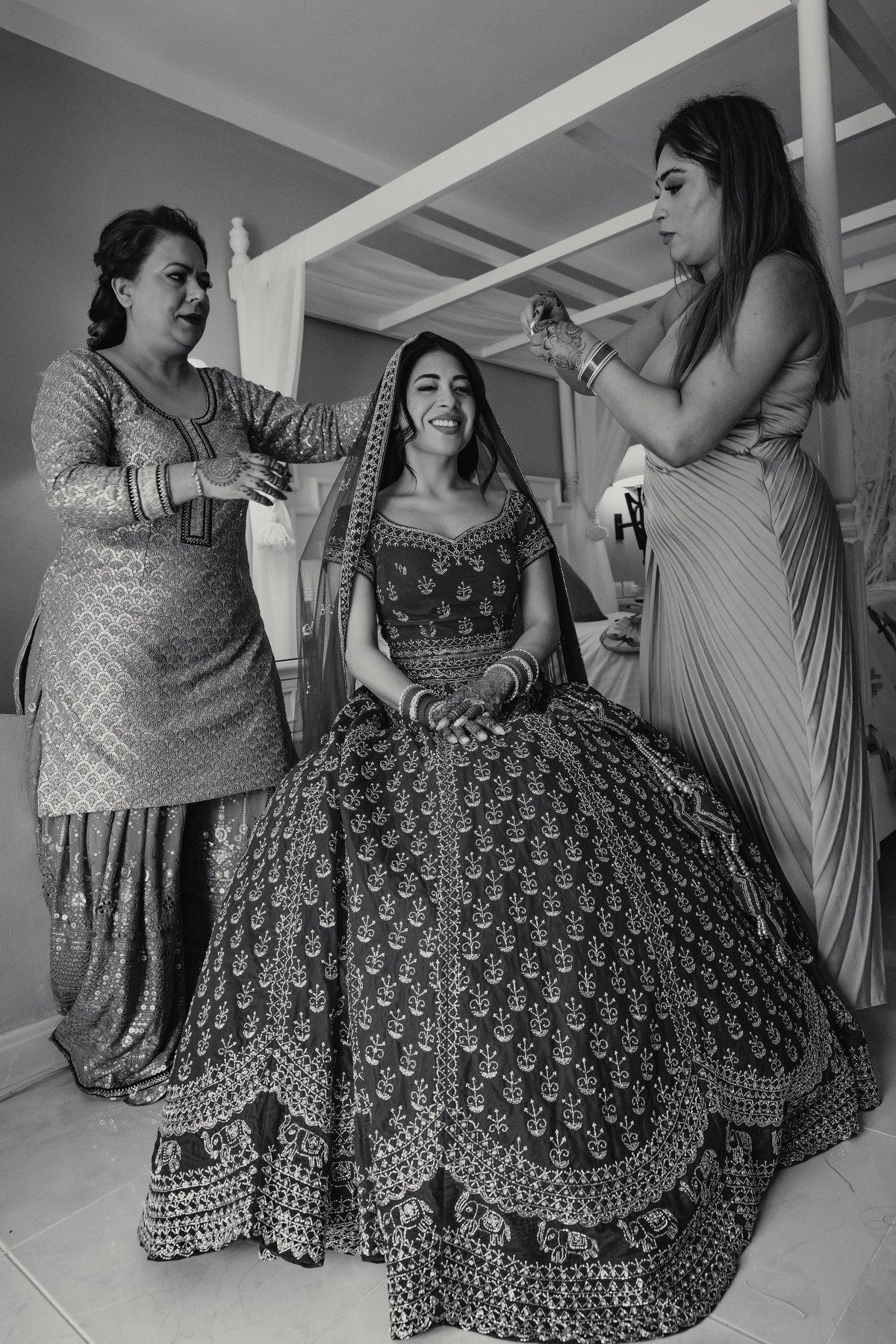 Bride in ornate lehenga laughing as two women help her prepare for Indian wedding ceremony.
