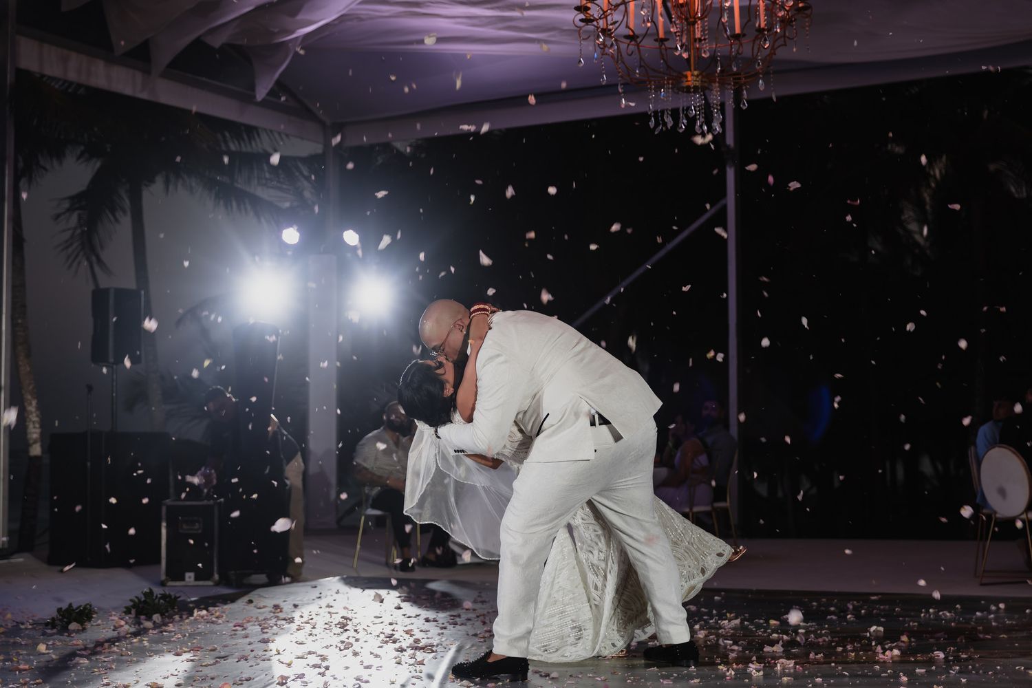 Groom dips bride in white gown during first dance as confetti falls at elegant evening wedding reception.