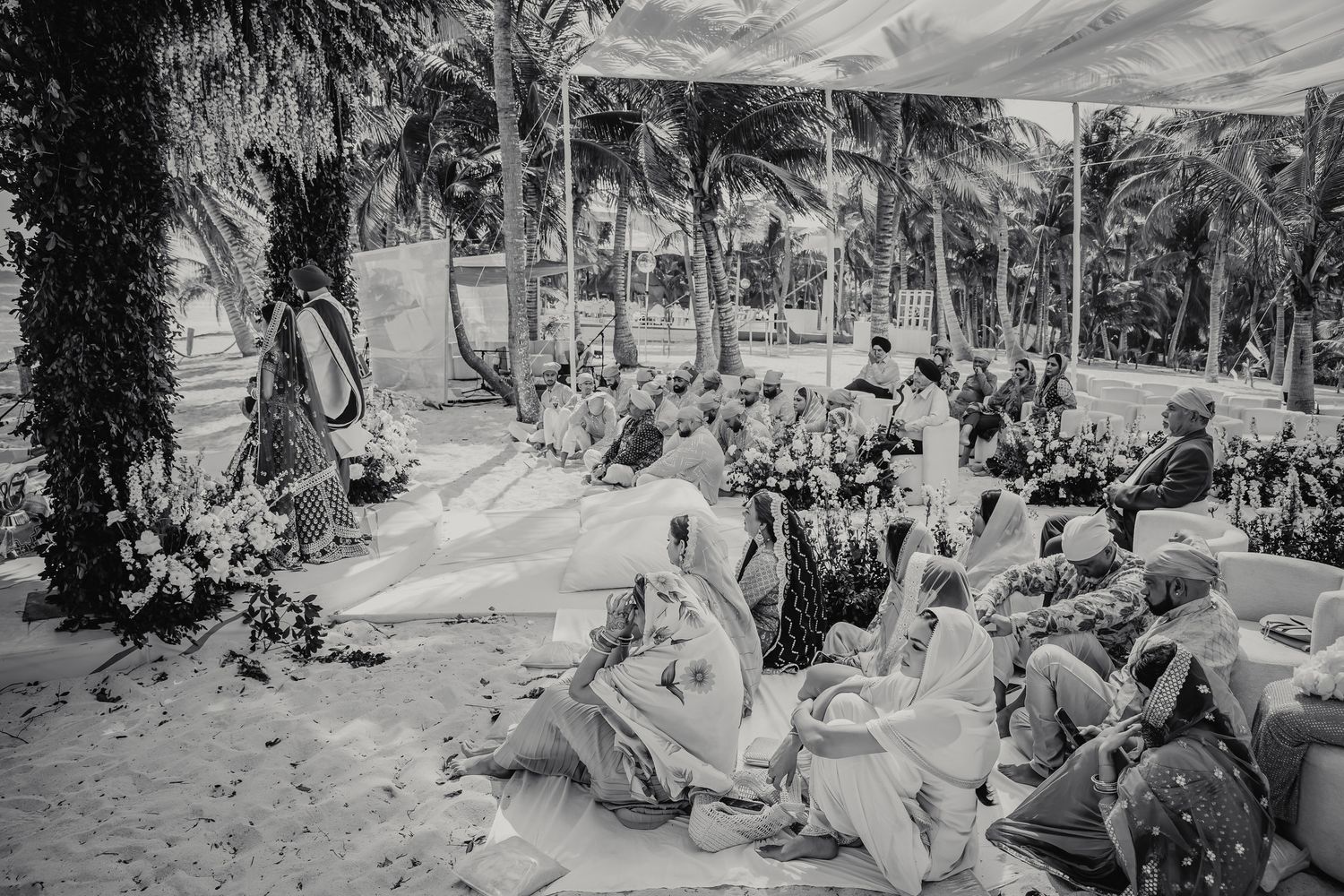 Black and white aerial view of a Sikh wedding ceremony on a sandy beach surrounded by palm trees.