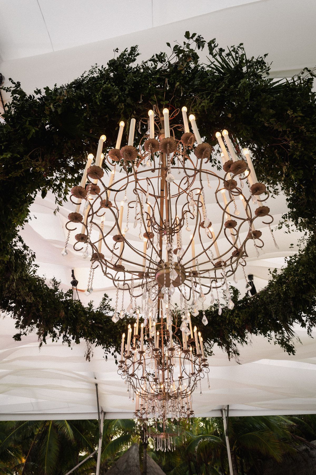 Elegant vintage crystal chandelier with candles encircled by a lush green wreath suspended from white wedding tent ceiling.