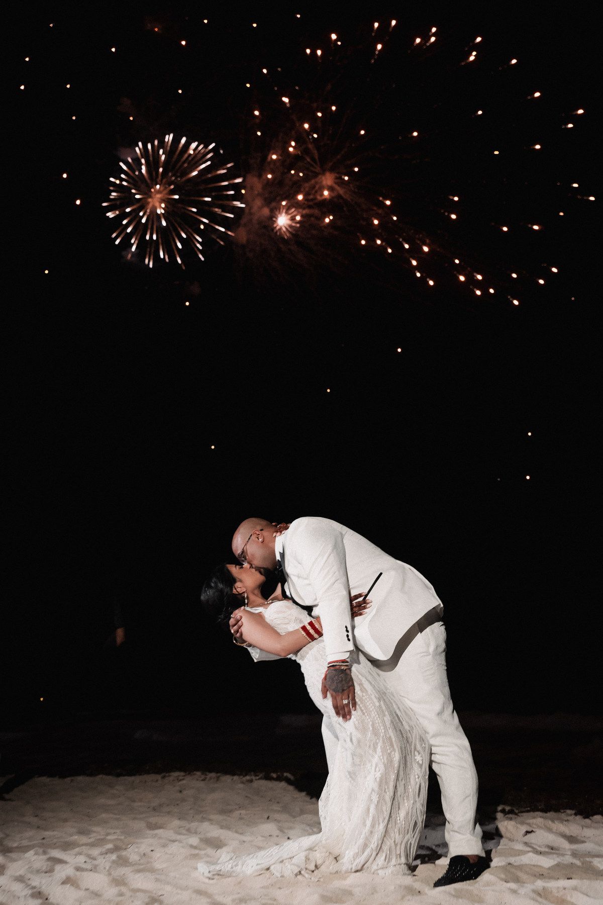 Couple sharing romantic dip kiss on beach with colorful fireworks display overhead.