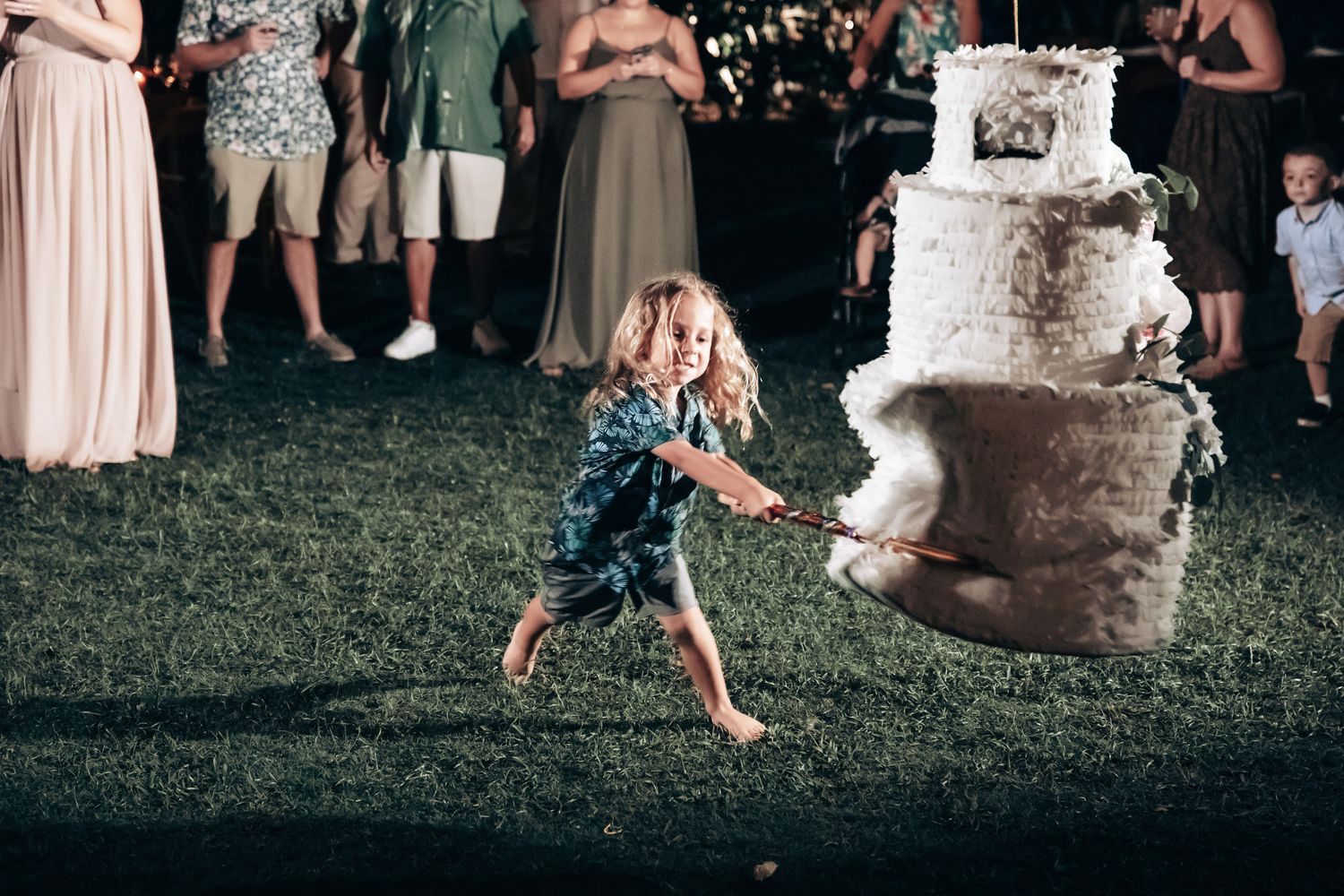 Young child pulls rope attached to wedding cake piñata at outdoor nighttime wedding celebration.