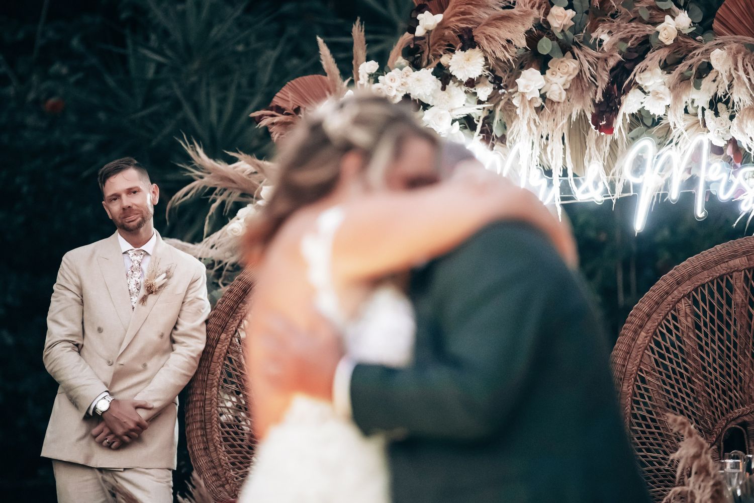 Groom watching emotional couple embrace near neon sign at boho wedding ceremony.